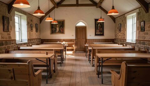 Small rural hall with wooden pews and tables arranged under warm pendant lights and stone walls